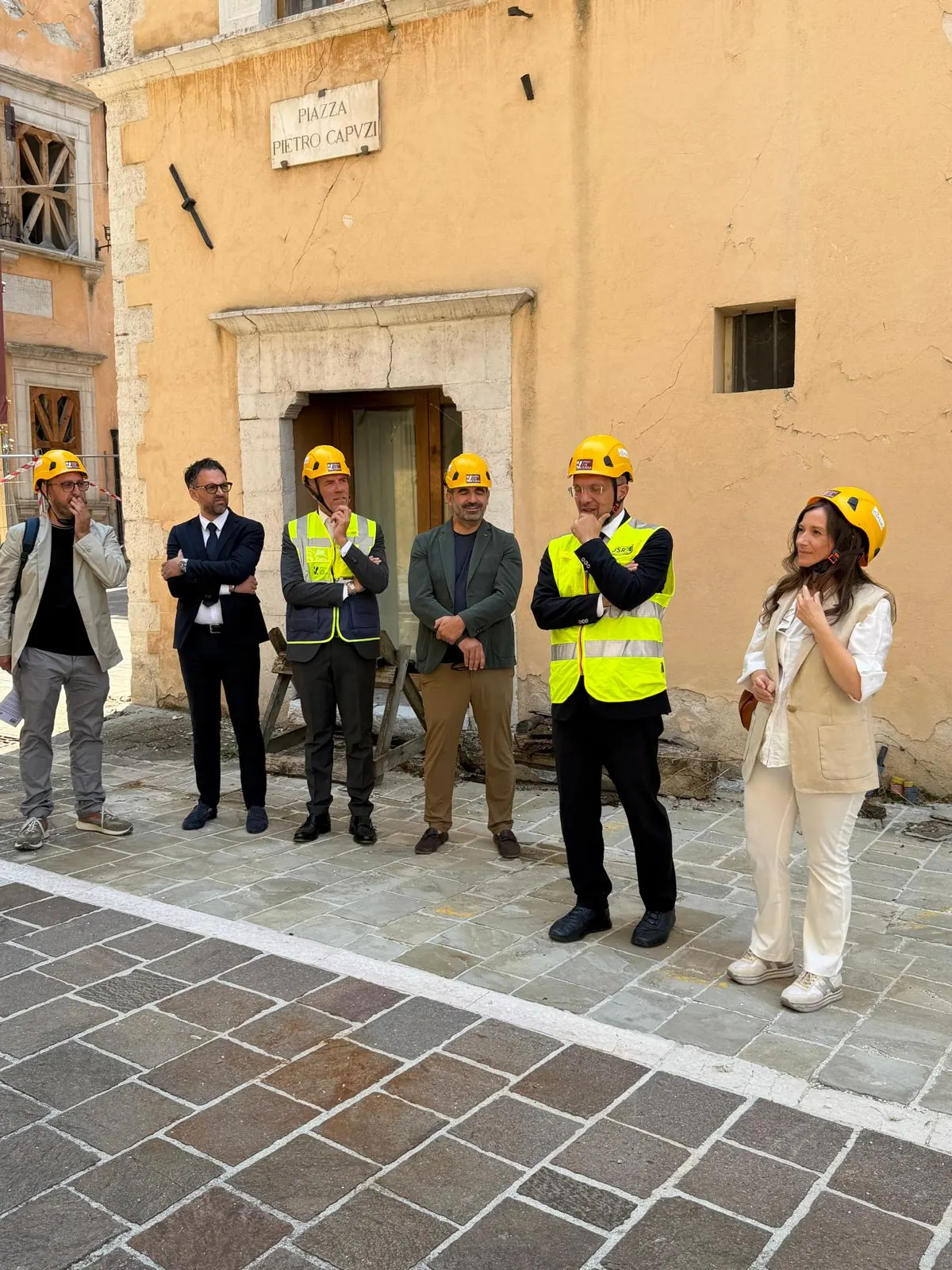 Gruppo di persone con elmetto giallo in piazza a Visso, davanti a un edificio storico
